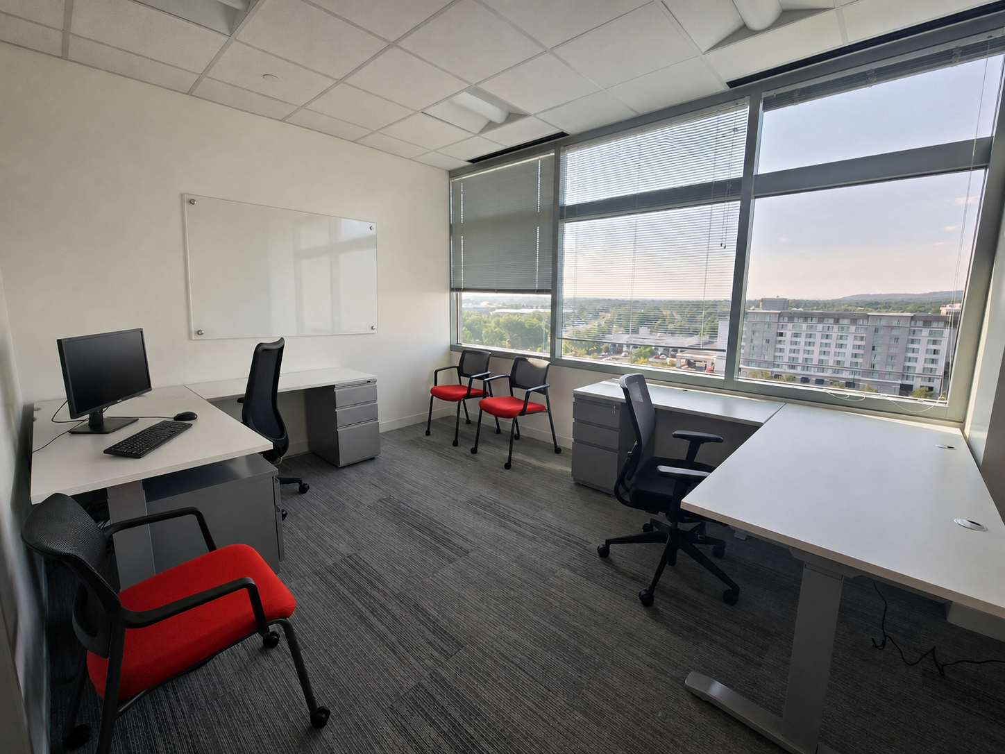 Modern private office with adjustable-height desk, dual monitor setup, red guest chairs, and floor-to-ceiling windows overlooking the city skyline